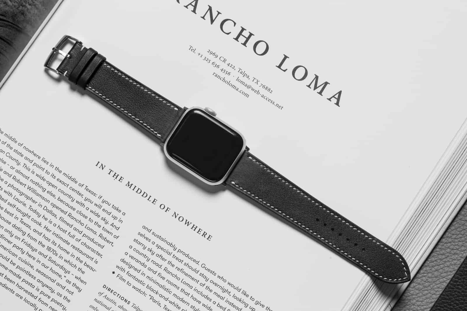 Black calf leather Apple Watch strap laid across an open book, showing the full band, the stainless adapter and the stitch line, manufactured by AKC
