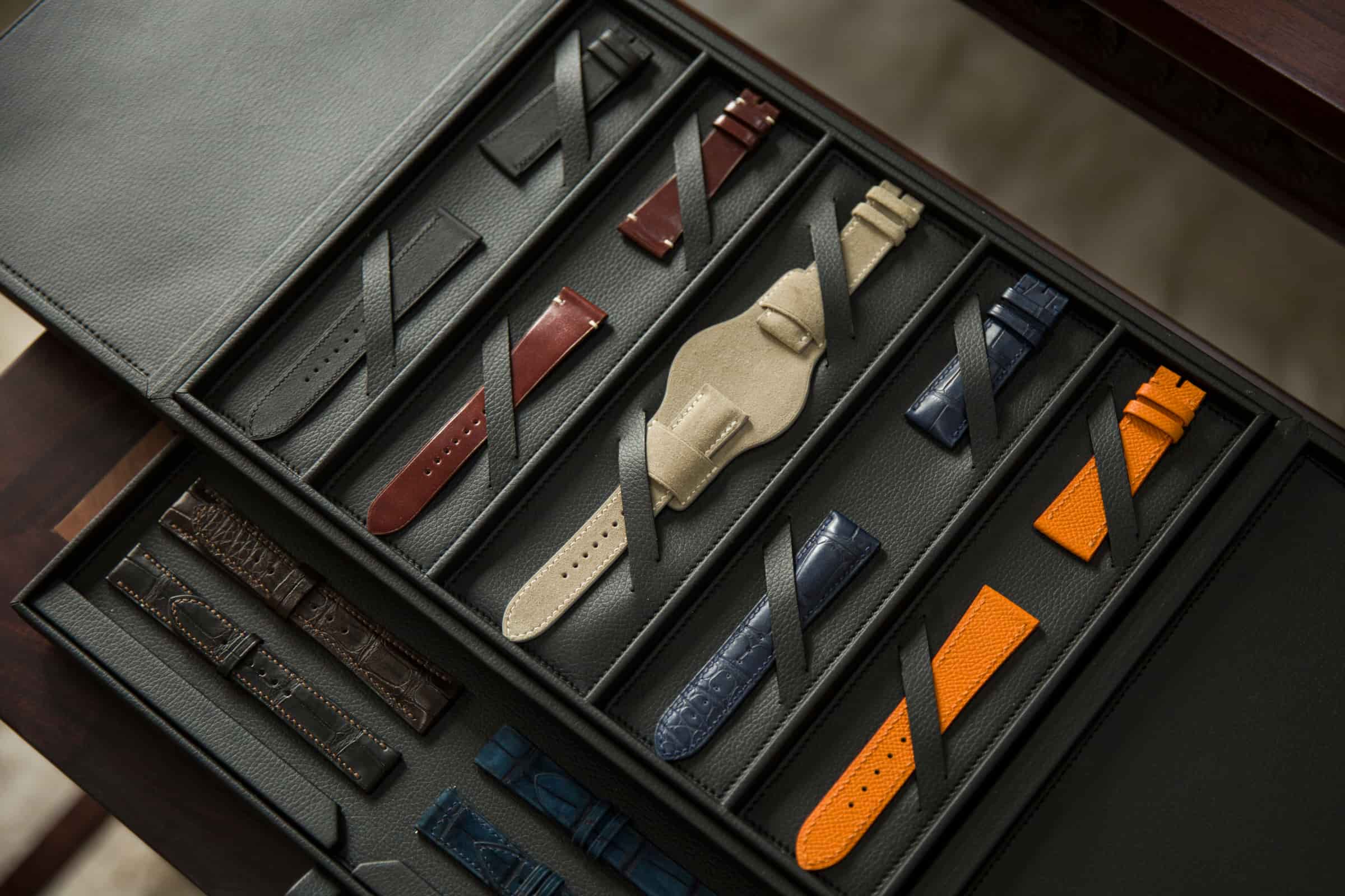 An open leather sample box holding two trays of watch straps in orange, red, navy, tan and black, on a workbench