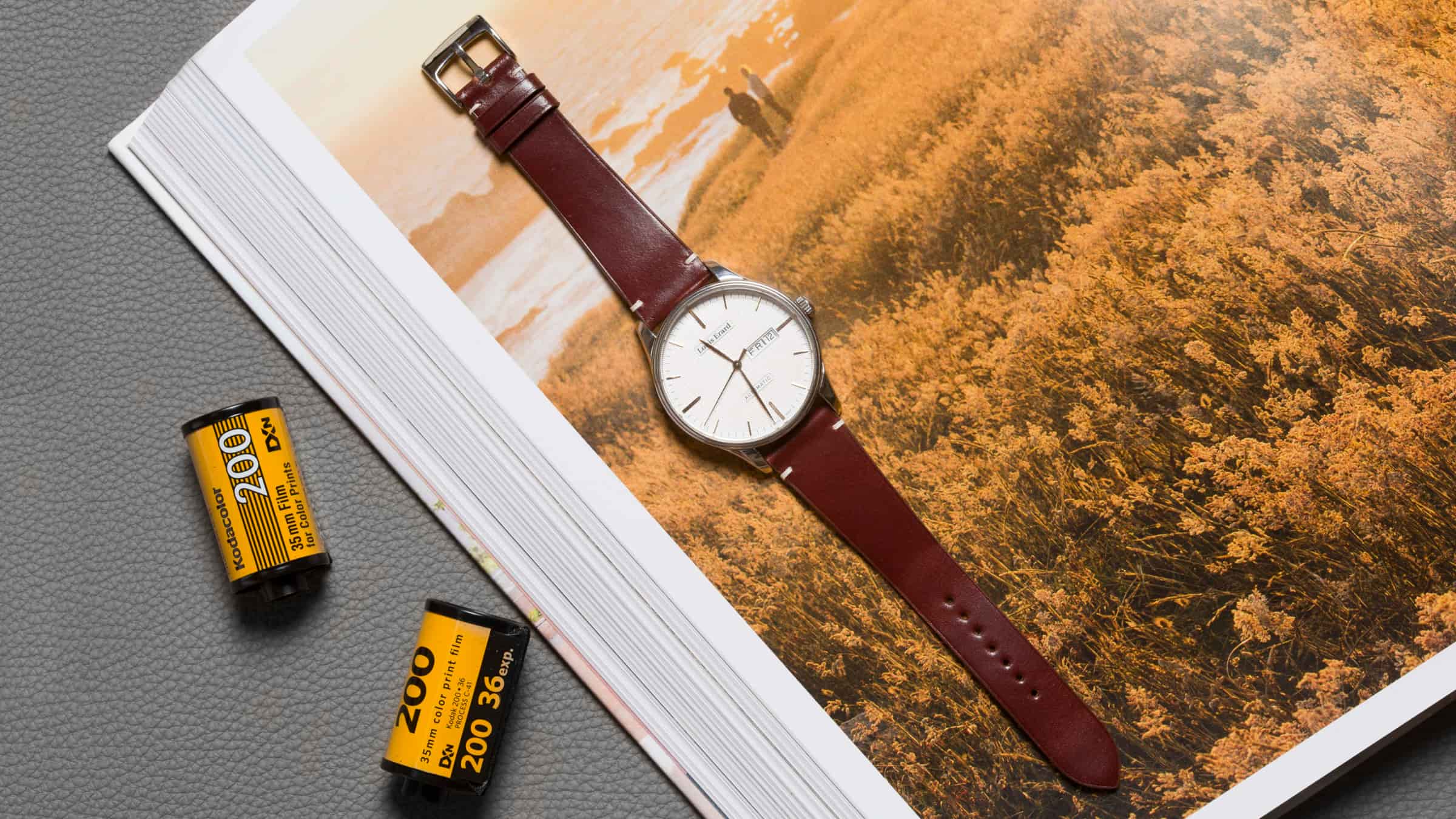 Brown calf leather watch strap on a white-dialled dress watch, lying on an open magazine spread of an autumn field, with two rolls of 35mm film beside it