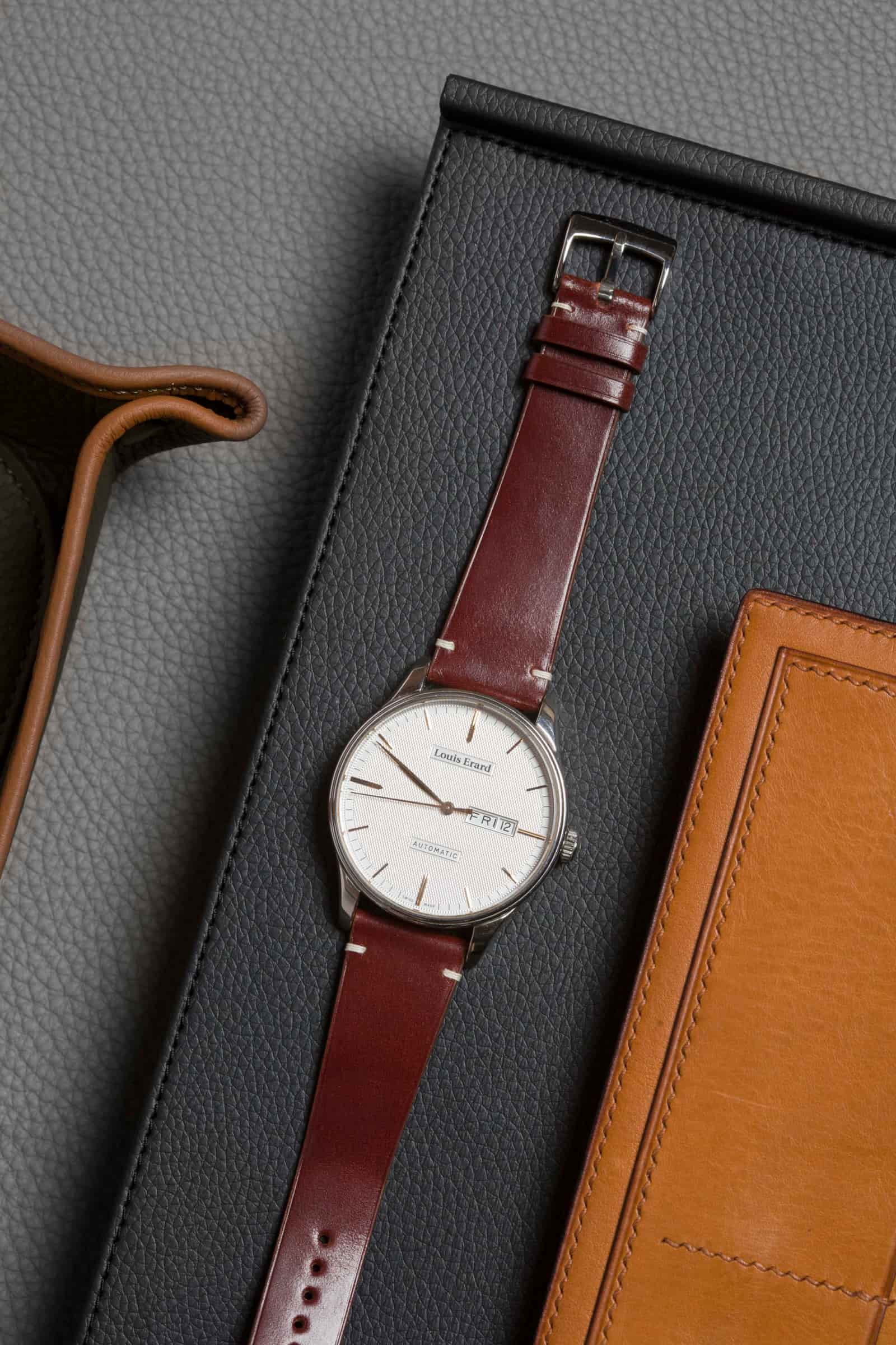 Oxblood calf leather watch strap on a white-dialled watch, laid on a grey pebbled-leather bag beside a wooden shoehorn and a tan leather card case