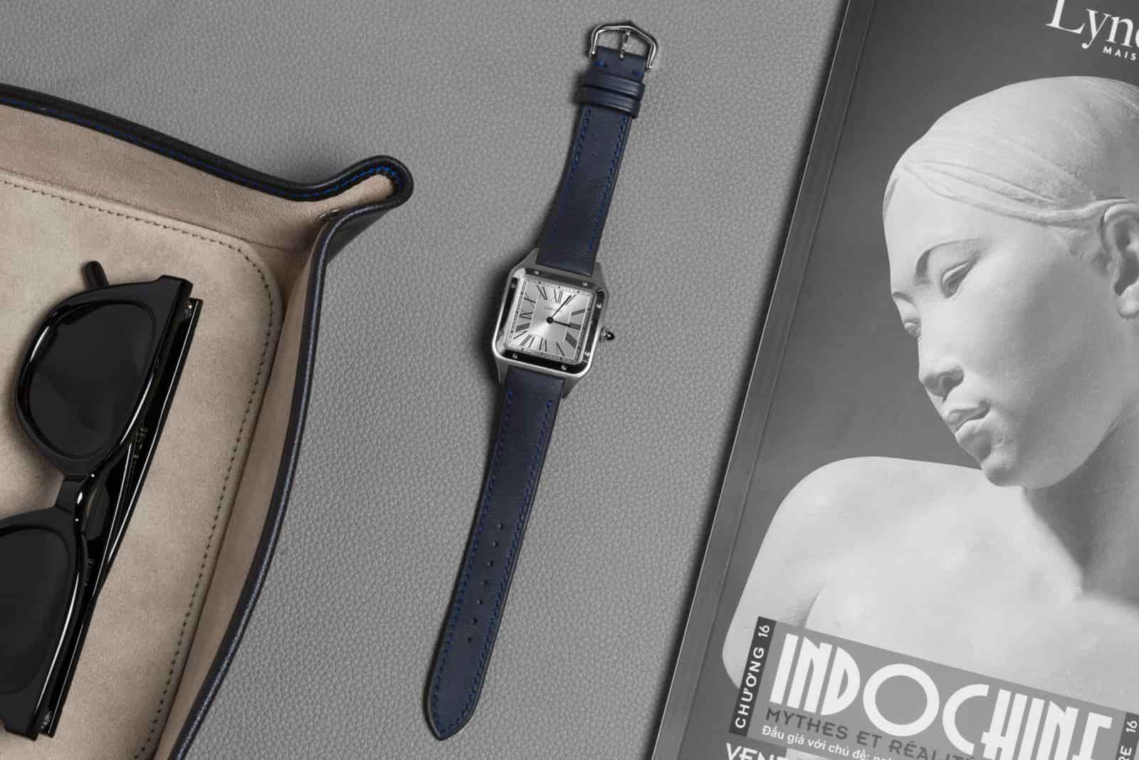 Navy smooth calf leather watch strap on a dress watch, staged on a desk beside sunglasses and an open magazine, made by AKC