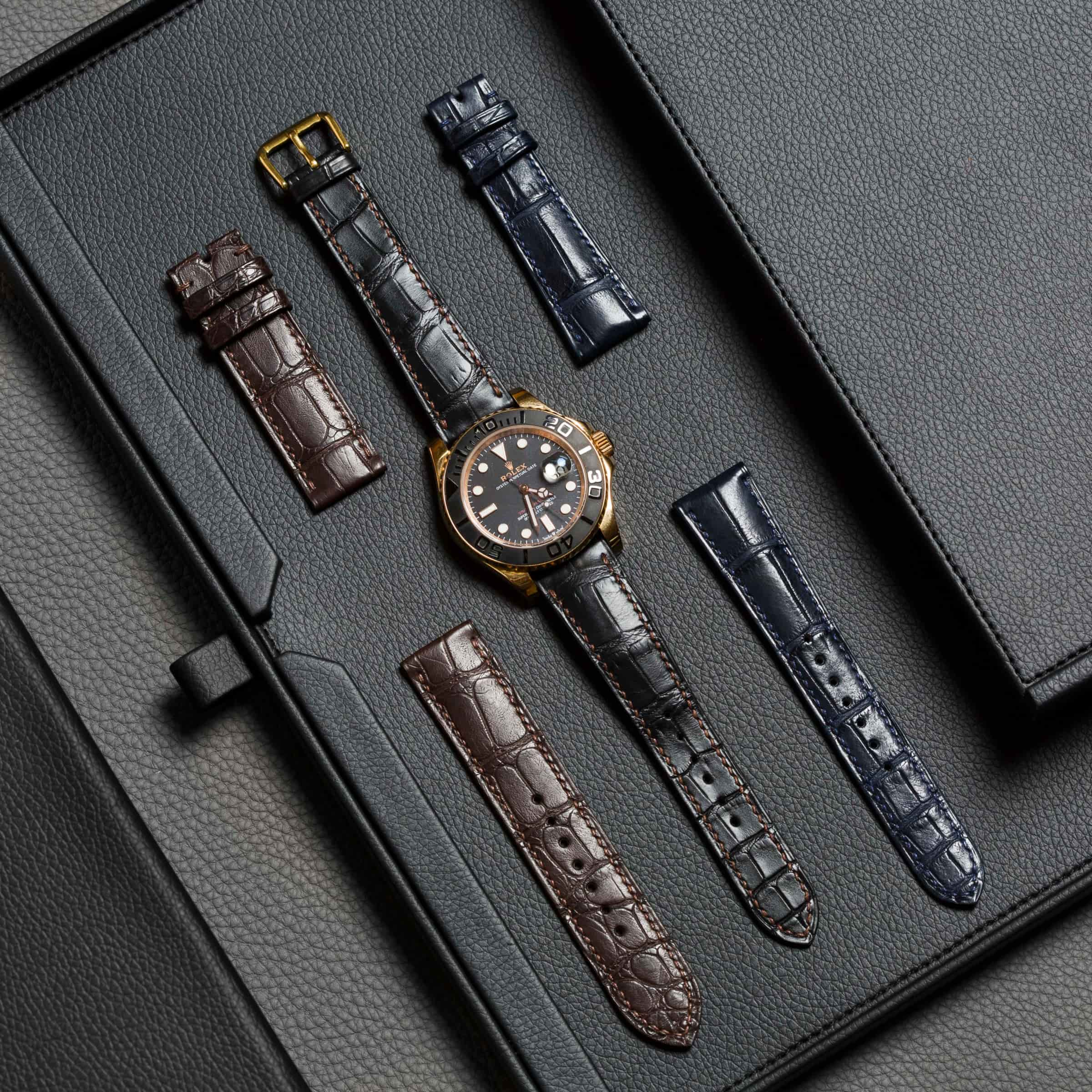 A gold-and-black dive watch on a black leather folio, ringed by five alligator watch straps in brown, grey and navy, each with its buckle fitted