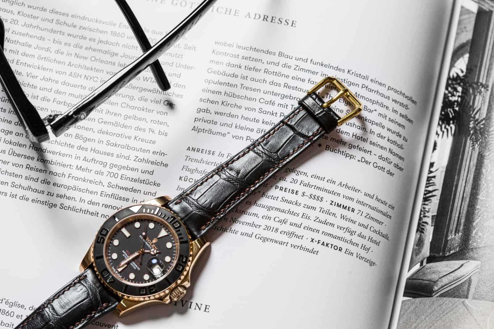 Grey alligator leather watch strap on a gold-and-black dive watch, lying across an open magazine spread with reading glasses behind it