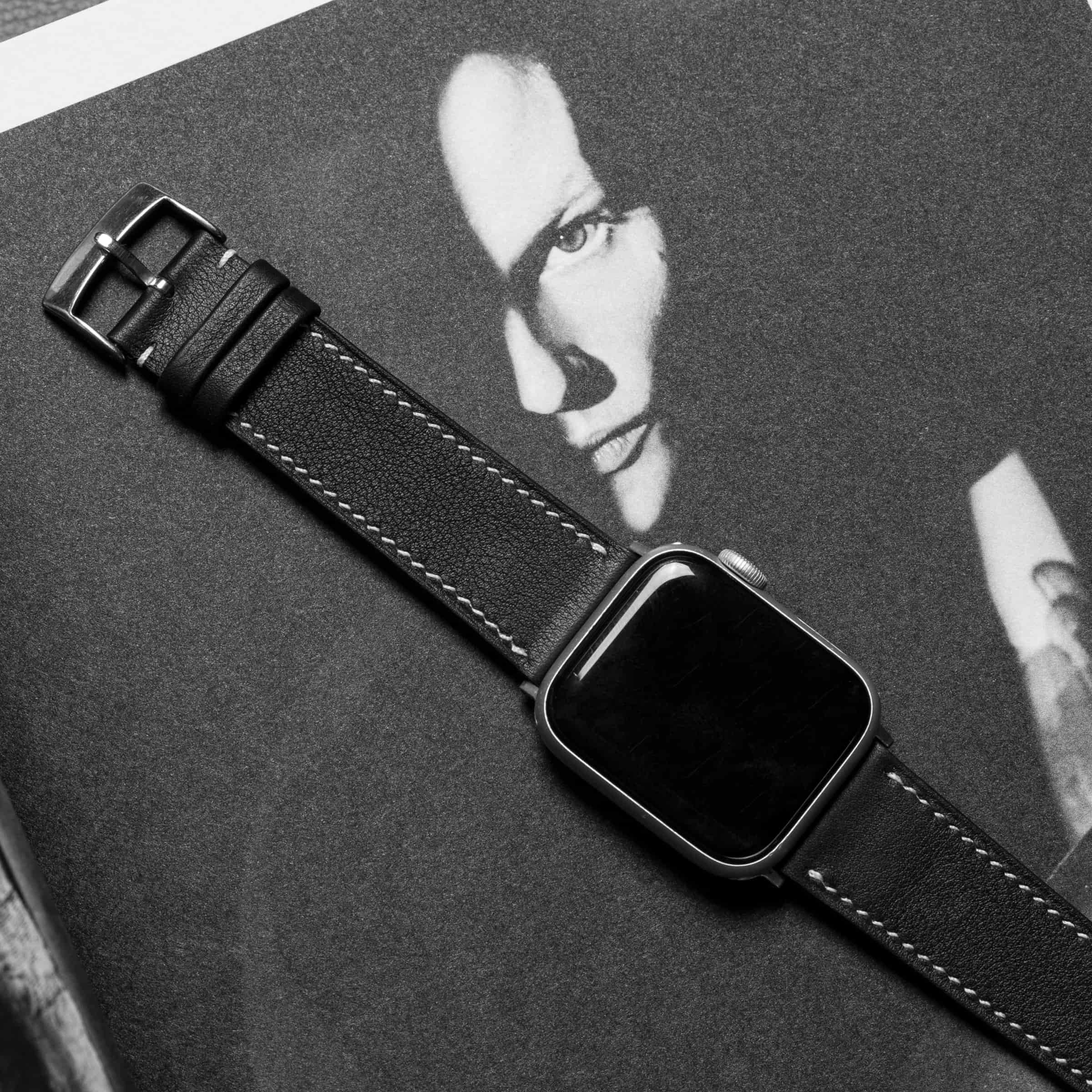 Black leather Apple Watch band with cream contrast stitching and a steel buckle, the watch fitted at its lower end, laid on grey beside a black-and-white portrait photograph