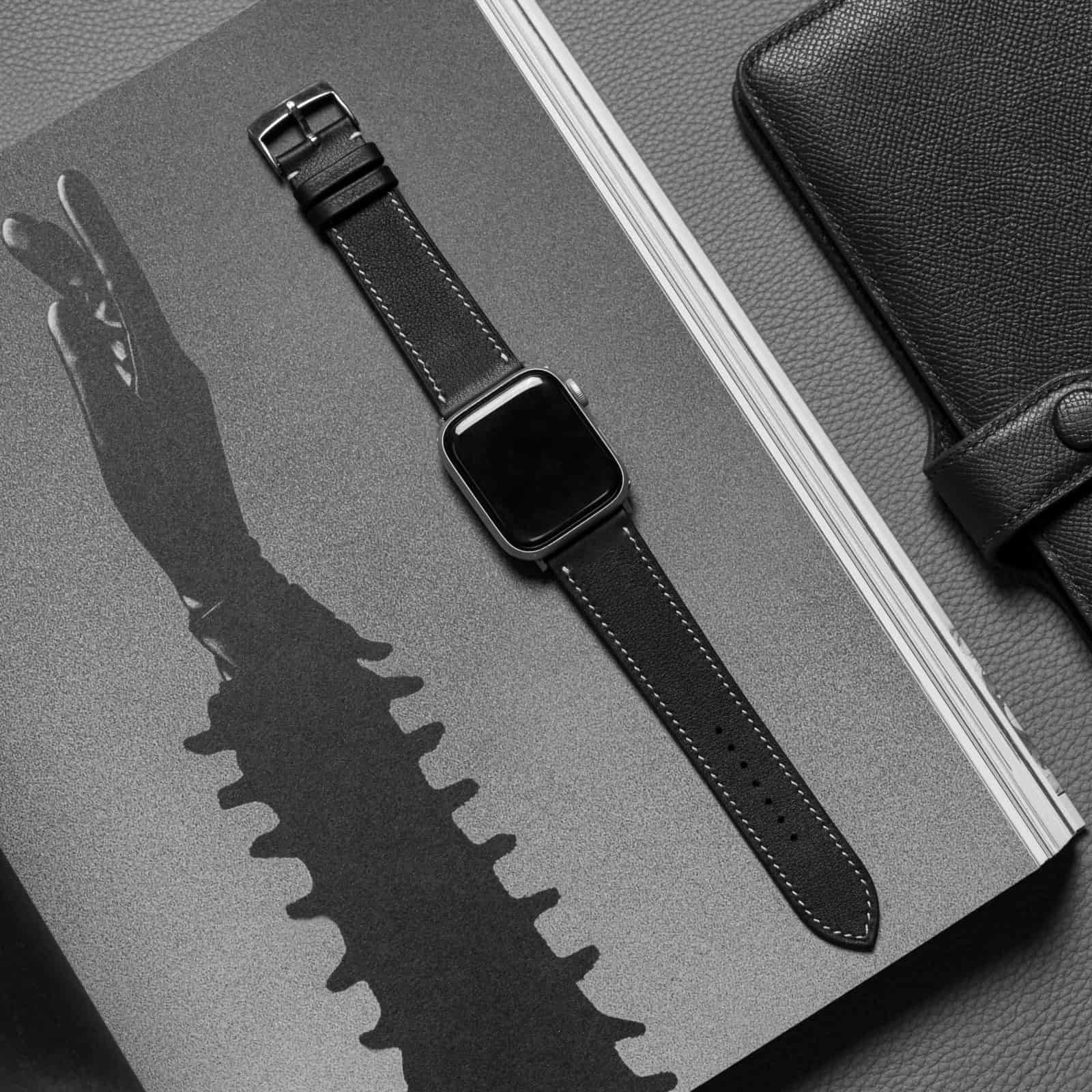 Apple Watch on a black leather band laid diagonally across grey, throwing its own shadow, the cream stitch line running the length of the band