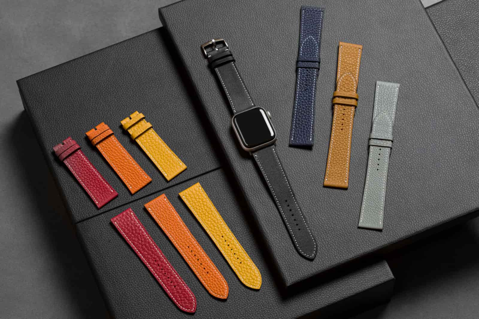 Leather Apple Watch bands in crimson, orange, yellow, navy, tan and grey laid across black leather folios around an Apple Watch fitted with a black band