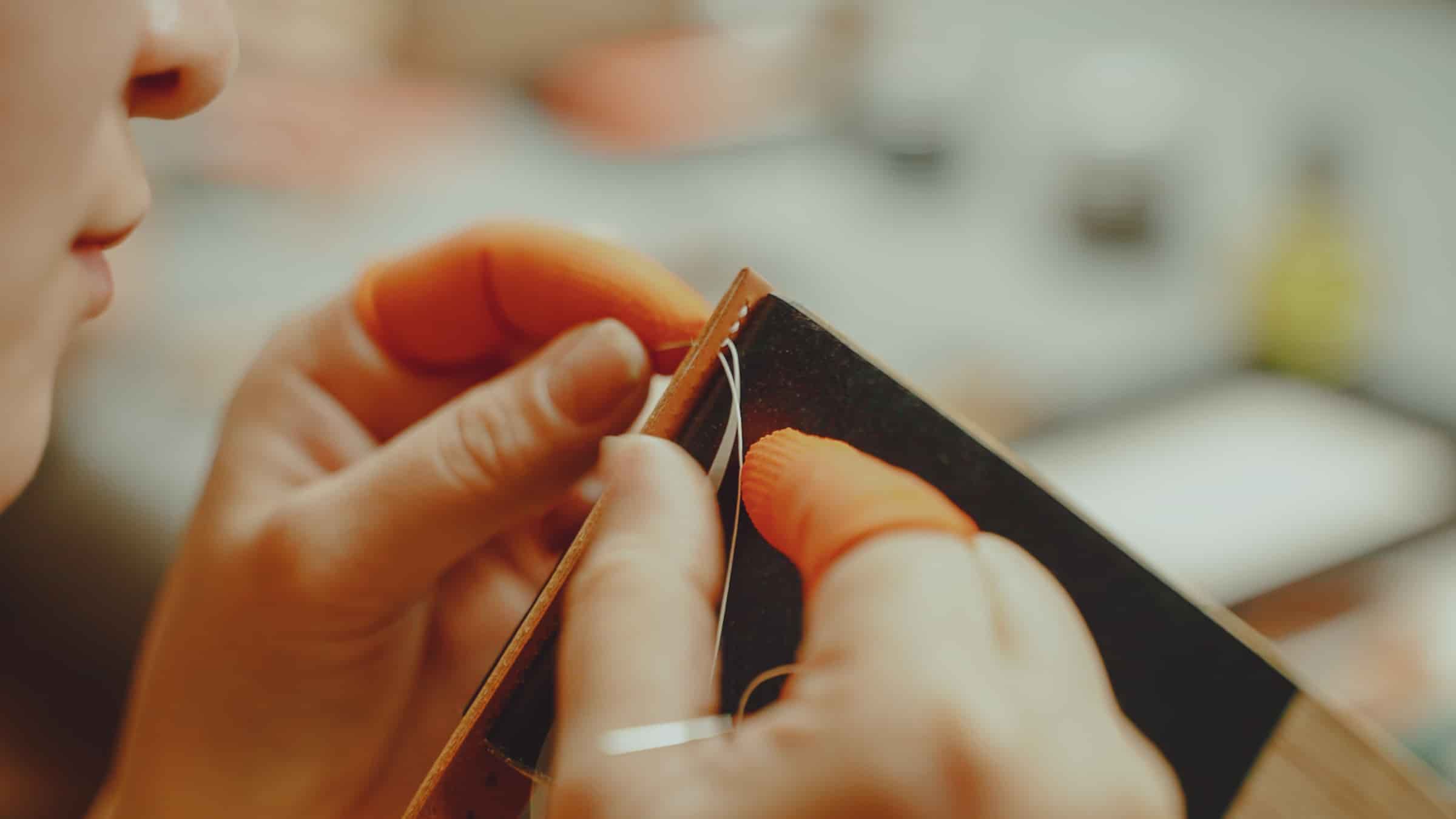 Two hands pulling a saddle stitch closed through an orange leather watch strap at the AKC bench