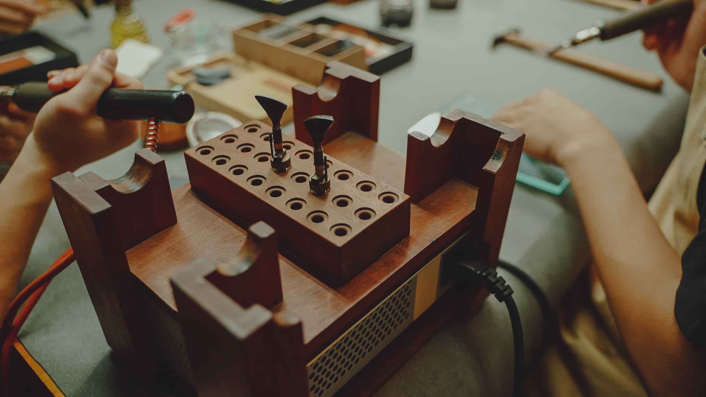 A heated edge-iron stand at the AKC bench, its wooden block holding a full rack of interchangeable creasing tips, with a craftsman's hands reaching in on either side