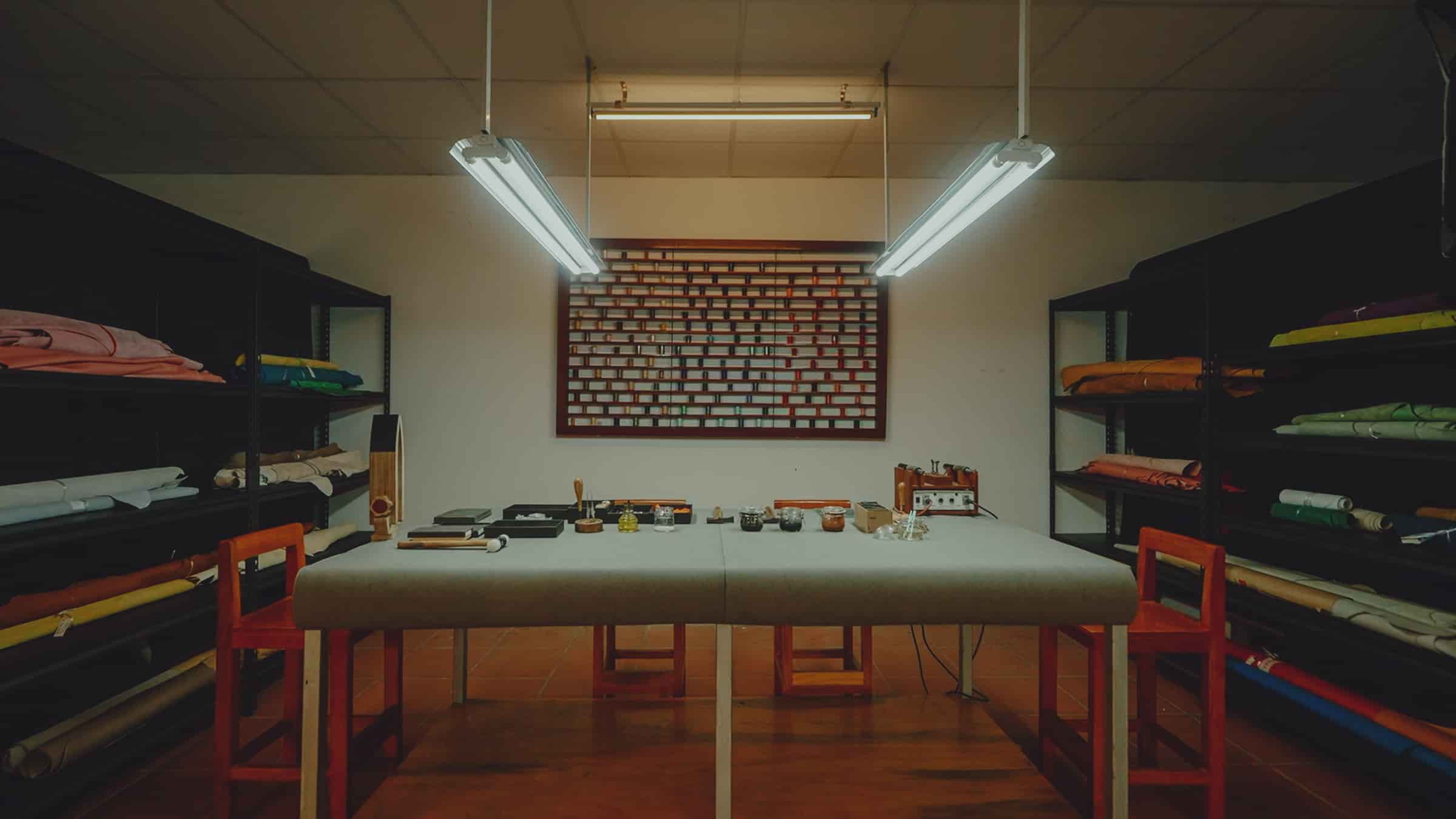 The AKC leather store in Vietnam: a long empty cutting table under strip lights, hides and rolls racked floor to ceiling on both walls, a board of leather swatches hung between them