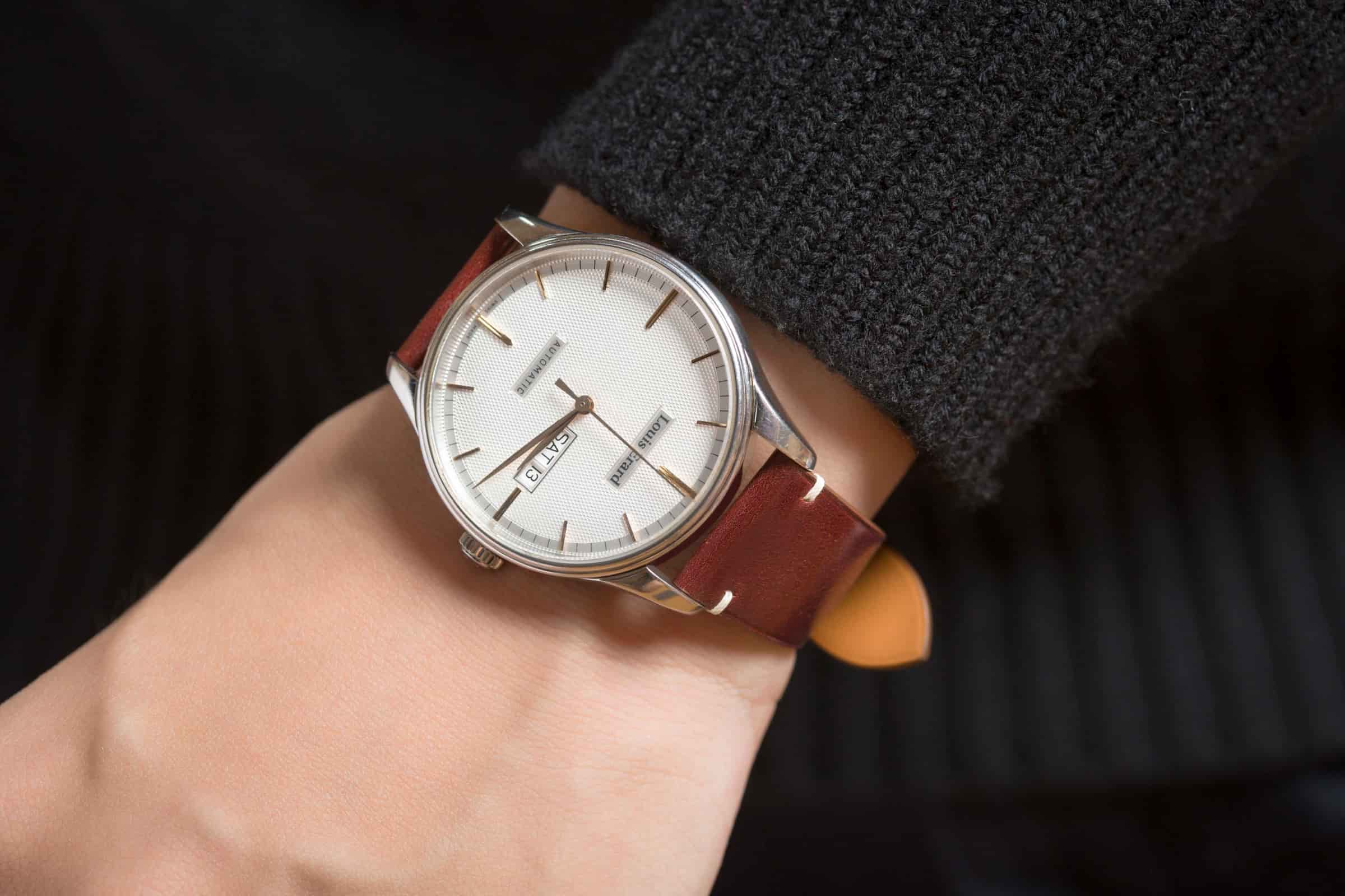 Grained brown calf leather watch strap on a black-dialled watch worn on the wrist against a tan background
